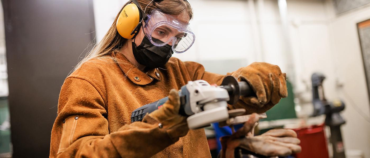 Student wearing protective equipment including goggles, headphones, mask, and gloves works in the sculpture studio.